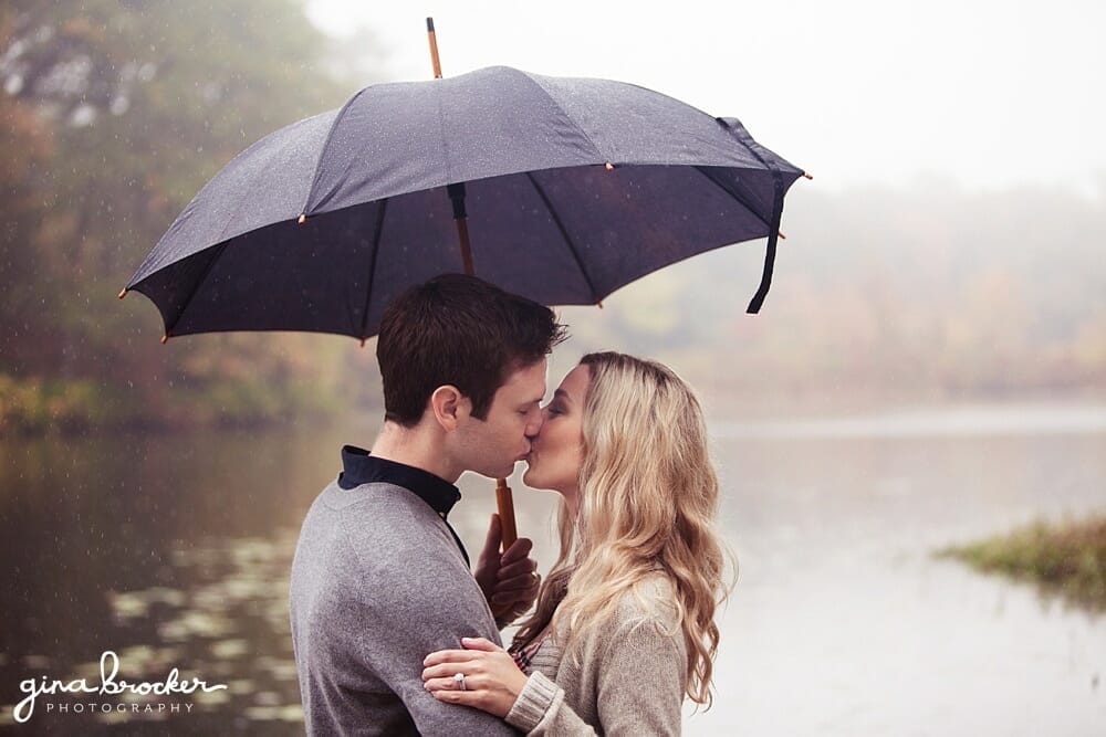 Engagement Session in the rain A couple hide under an umbrella and kiss during their rainy engagement session on the charles river in boston massachusetts