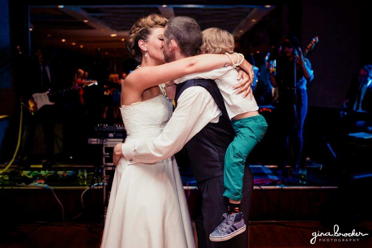 The bride and groom hug and kiss while they dance with their son during their fun and retro wedding