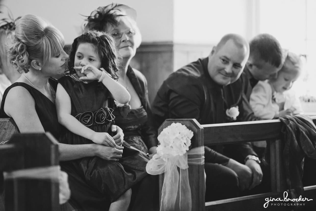 A candid photograph of the flower girl sitting on her mothers lap during a church wedding ceremony