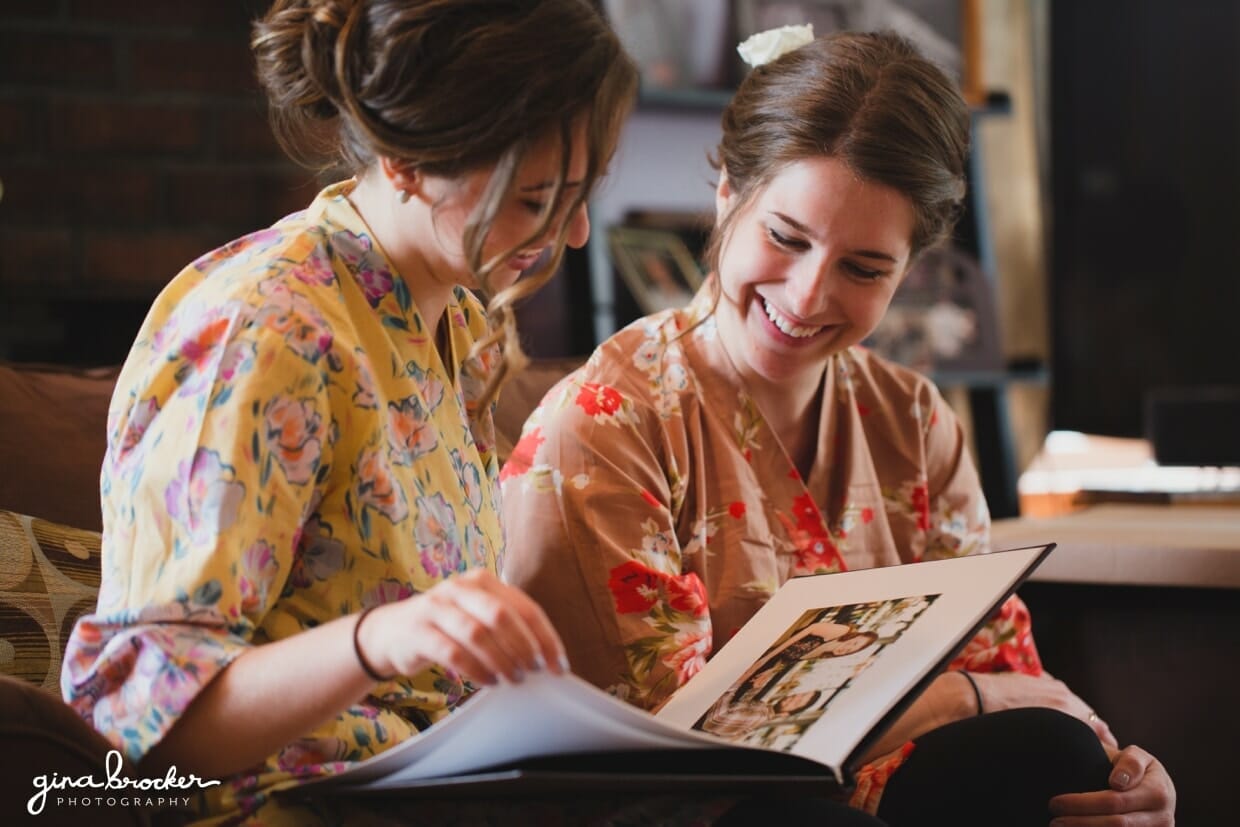 Two bridesmaids wearing floral robes look at the photo guest book of the bride and groom on the morning of their wedding