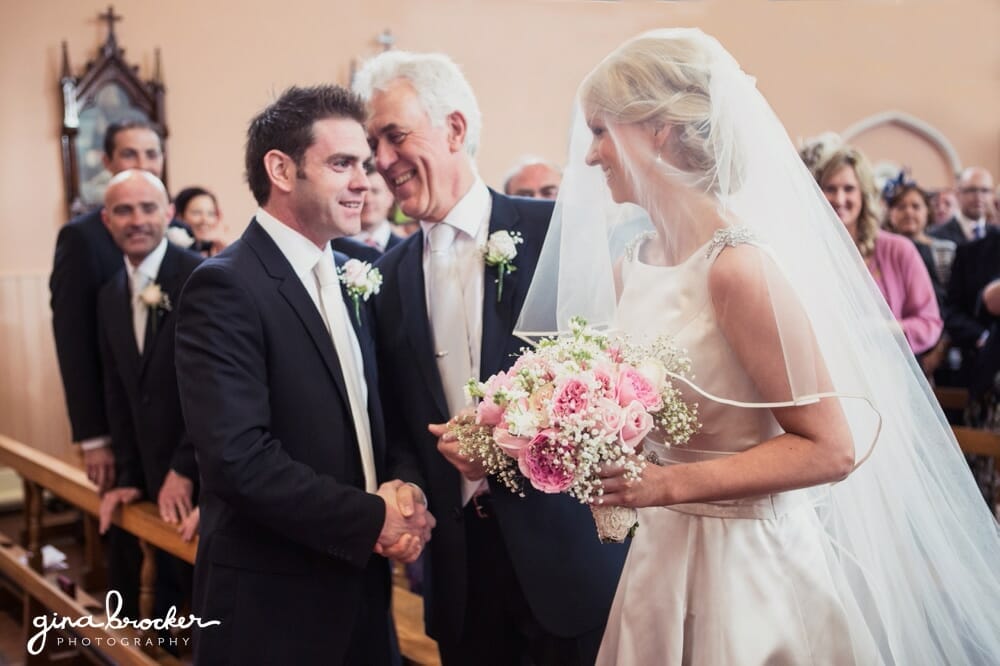 A sweet photograph of a groom shaking the father of the bride's hand as he stares lovingly at his bride during their church ceremony in Boston, Massachusetts
