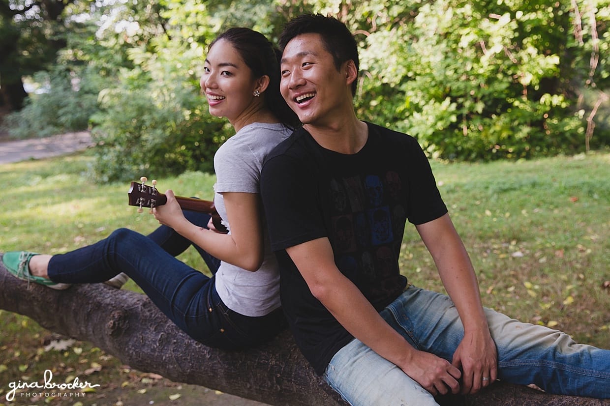 A couple laugh together as they sit on a tree and play music during their Boston Engagement session in Back Bay Fens