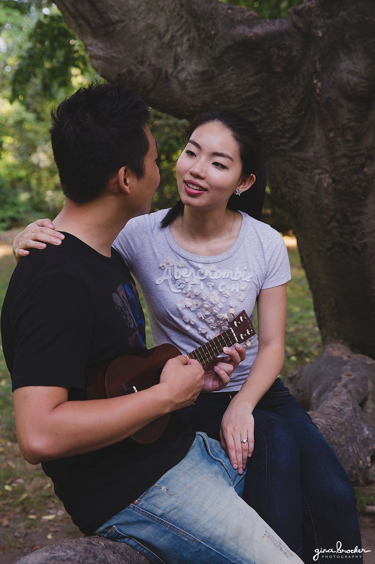 A couple sit on a tree and play music in Back Bay Fens during their park engagement session in Boston, Massachusetts
