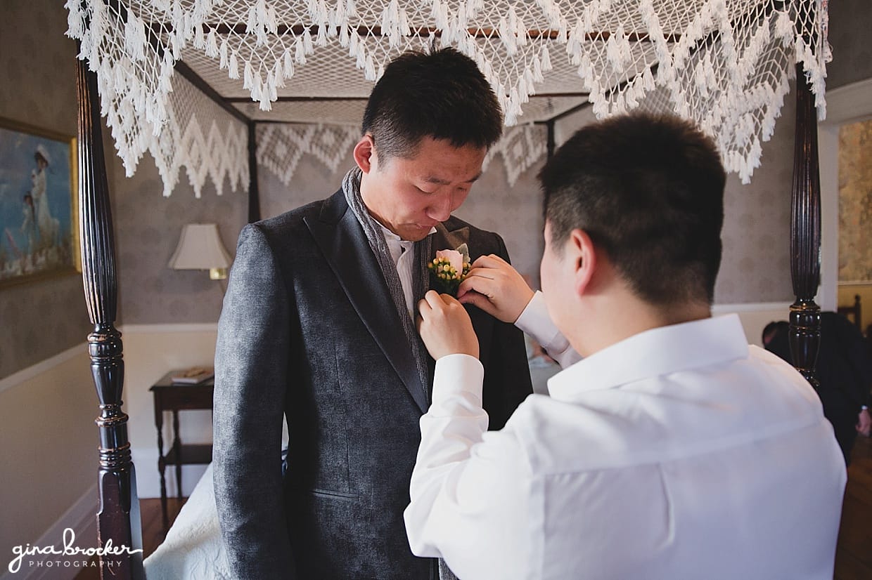 A candid photograph of a groomsmen pinning a boutonnière on the groom on the morning of his hammond castle wedding in gloucester, massachusetts