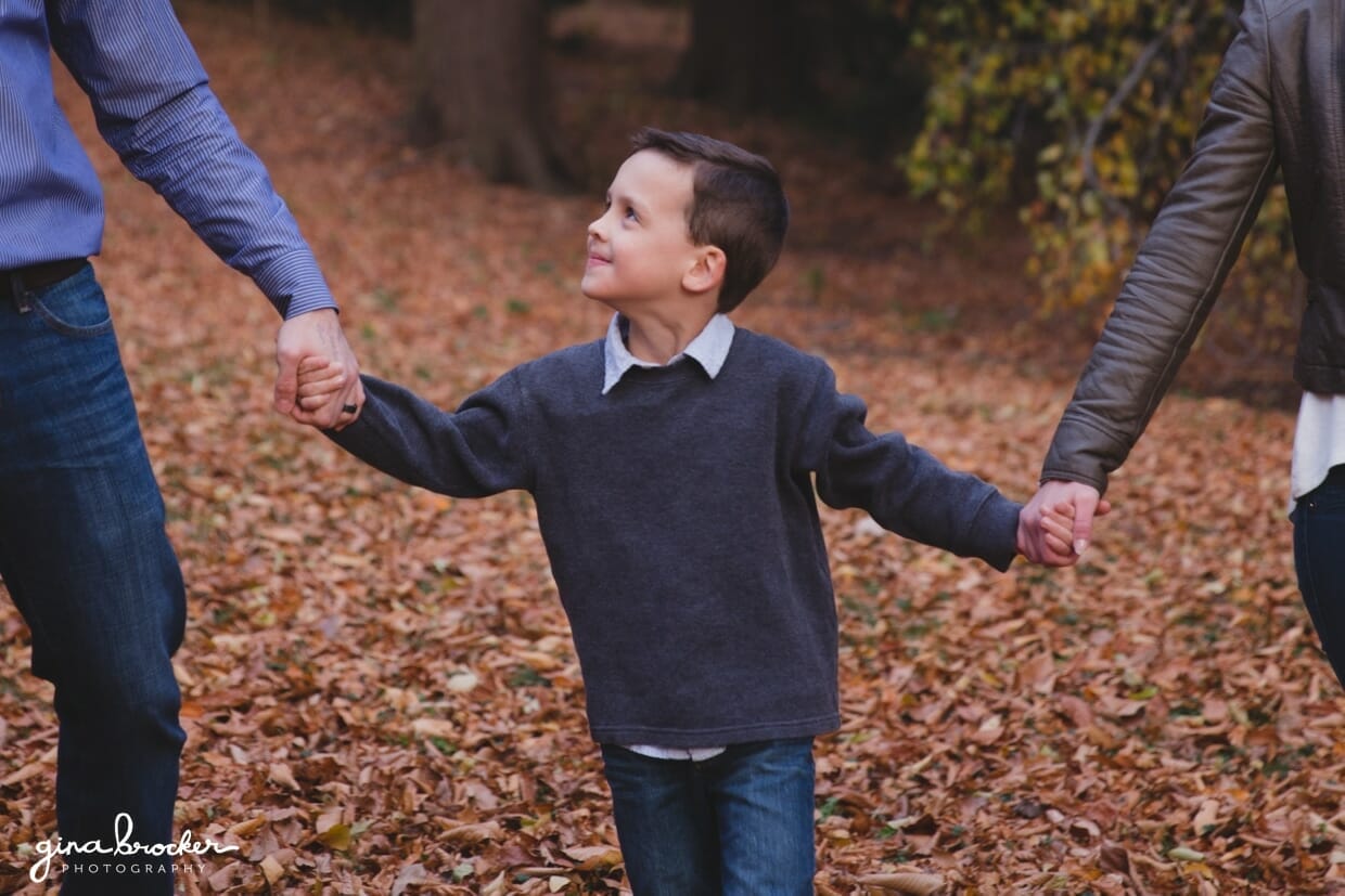 A sweet photograph of a son looking up at his father as they walk together in the woods during their fall family photo session at the Arnold Arboretum in Boston, Massachusetts