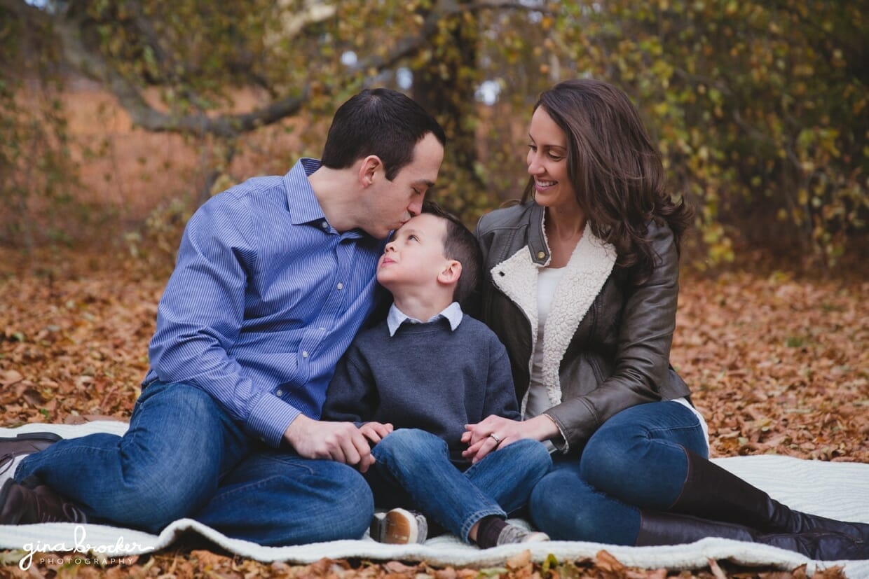 A dad gives his son a kiss on the forehead while he and his family sit on a blanket in the woods during a fall family photo session in Arnold Arboretum