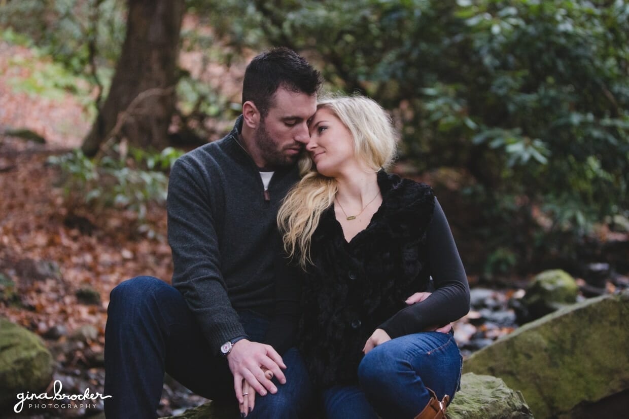 A sweet photograph of a couple sitting together on a rock in the woods during their fall couple session at the Arnold Arboretum in Boston, Massachusetts