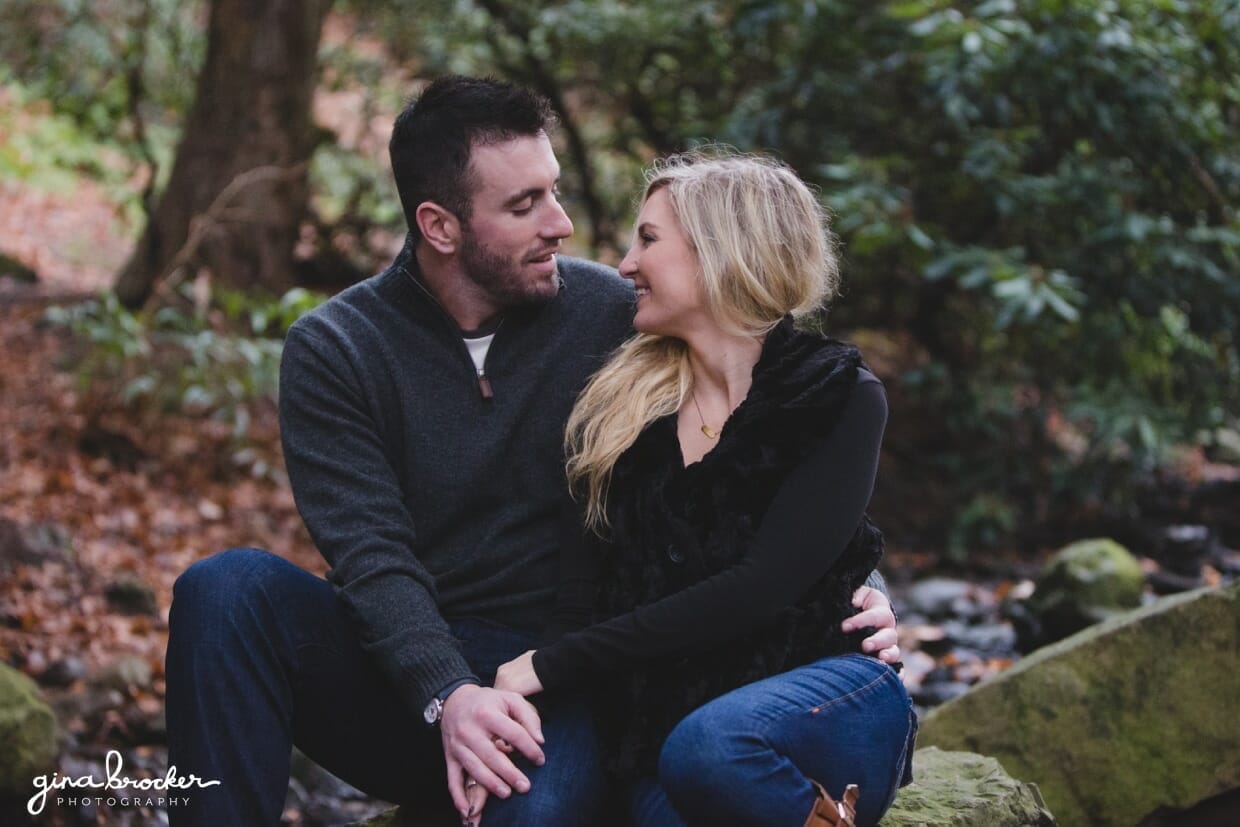 A natural and sweet photograph of a couple laughing together as they sit on a rock in the woods during their fall couple session in Boston's Arnold Arboretum