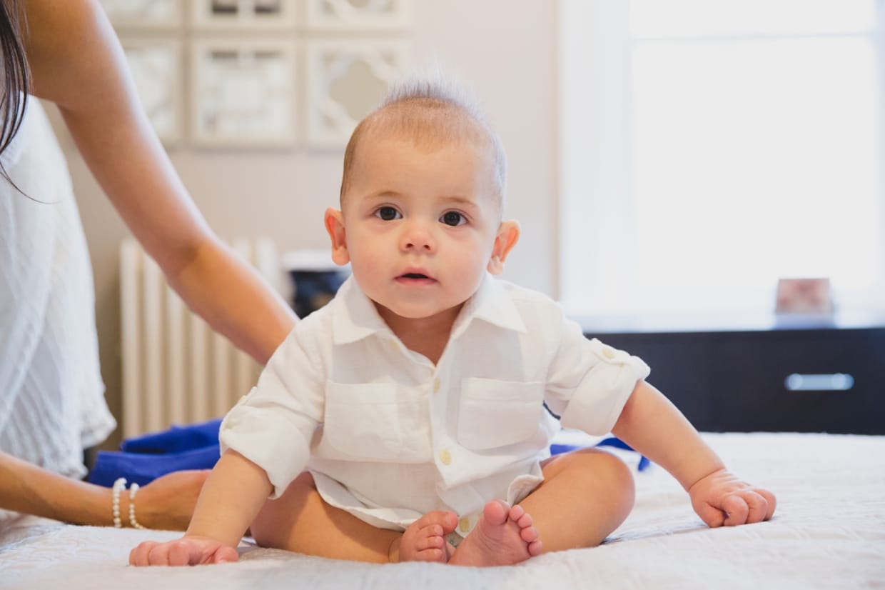 A sweet portrait of a baby boy sitting on the bed while he gets dressed for a family photo session in his Boston home.