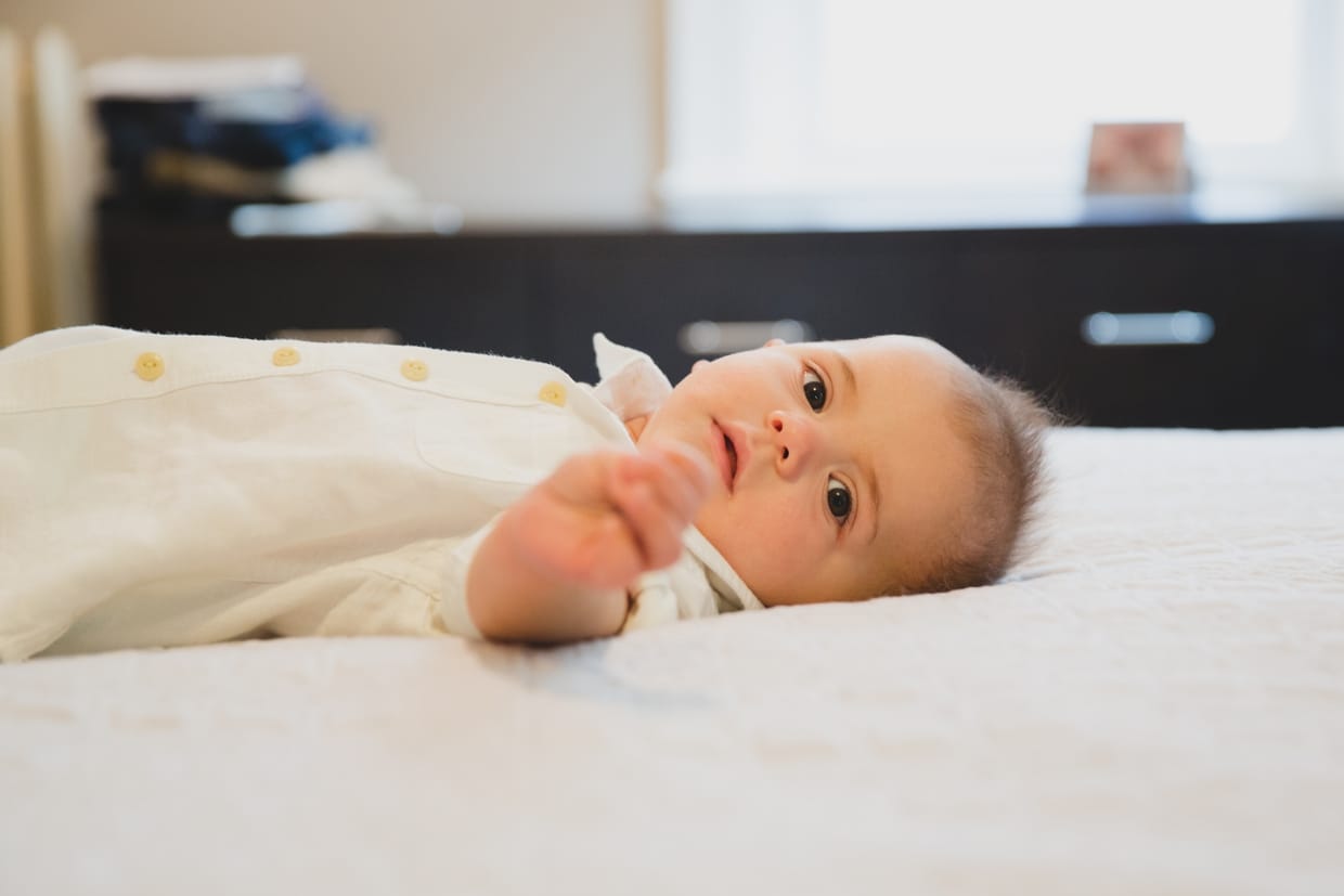 A beautiful natural portrait of a baby boy laying on the bed while he gets dressed for a family photo session in his Boston home.