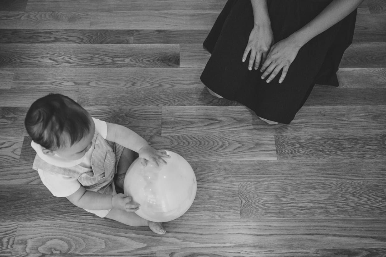 An artistic and documentary photograph of mother watching her baby boy play with a ball during their in home family photo session in Jamaica Plain