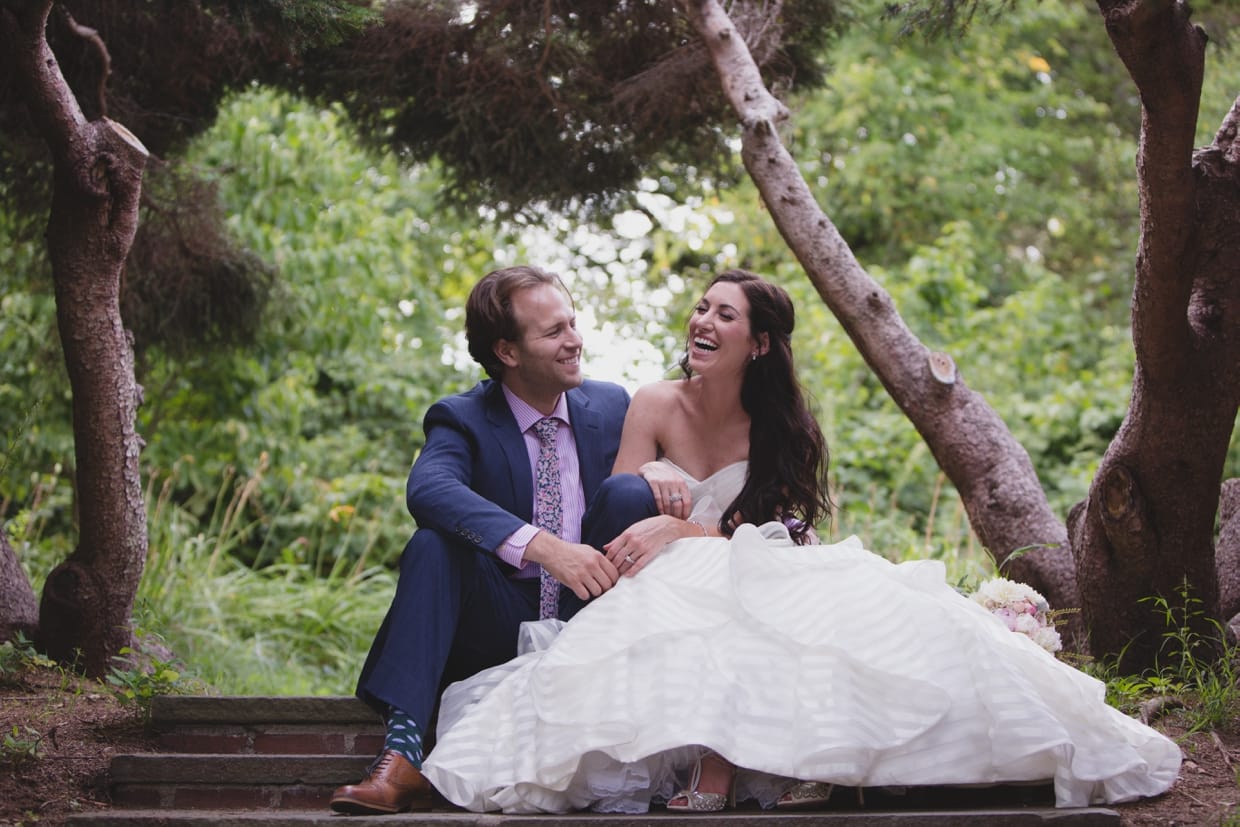 A sweet wedding portrait of a couple laughing as they sit on the steps of the Minute Man Park during their first look