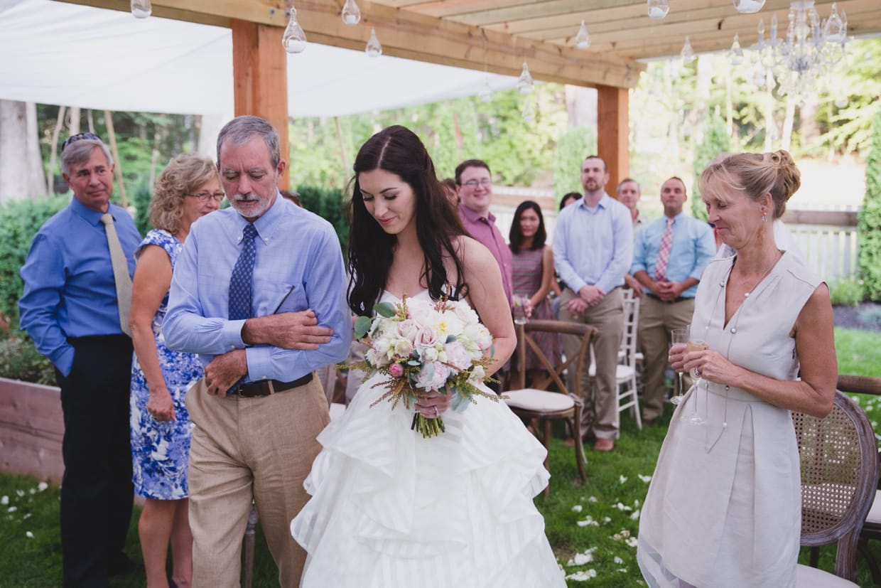 Guests smile as a father walks his daughter down the aisle during a backyard wedding ceremony in Massachusetts