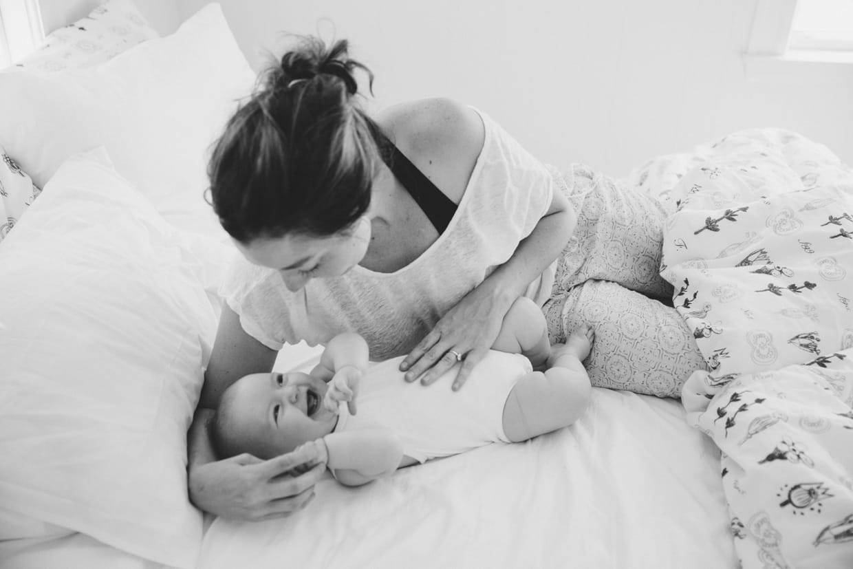 A beautiful photograph of a baby laughing while laying in bed with her mother during a Boston in home family photo session