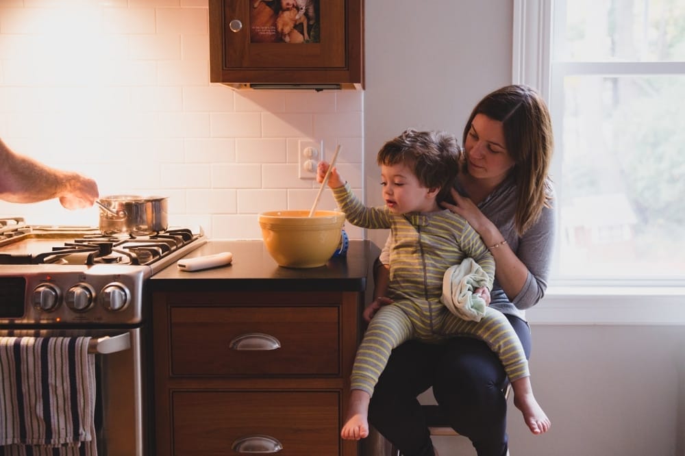 A documentary photograph of a little boy stirring pancake batter while sitting on his mom's lap during their lifestyle family photo session at home in Boston, Massachusetts