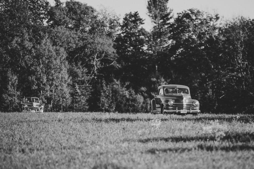A documentary photograph of two wedding cars driving up a grassy hill during a rustic outdoor wedding ceremony in New Hampshire at Kitz Farm