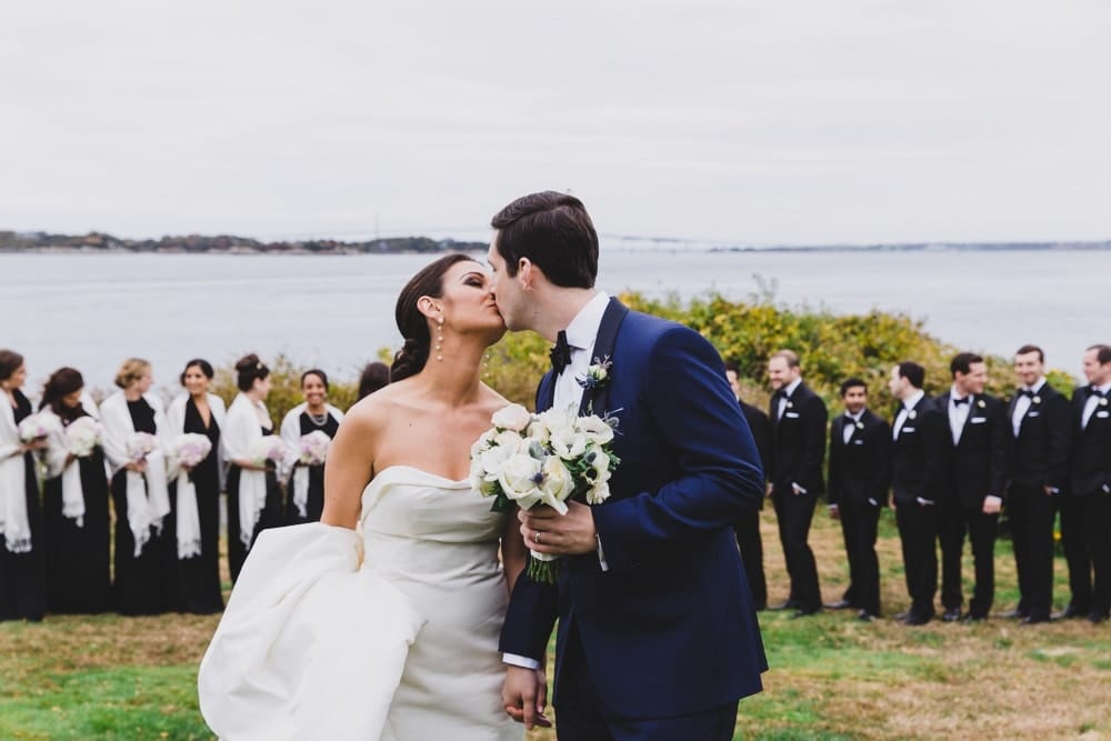 A portrait of a bride and groom kissing with their wedding party in the background at the Castle Hill Inn in Newport, Rhode Island