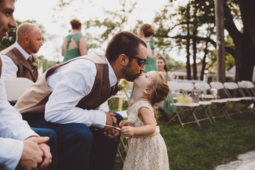 A photojournalistic photograph of a groomsman kissing his daughter during a rustic outdoor River Club Wedding in Scituate, Massachusetts