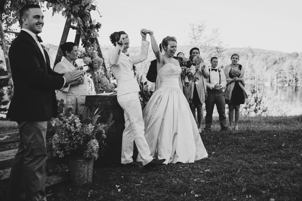 A documentary photograph of two brides celebrating as they walk down the aisle as wife and wife after their rustic outdoor wedding ceremony at New Hampshire's Kitz Farm