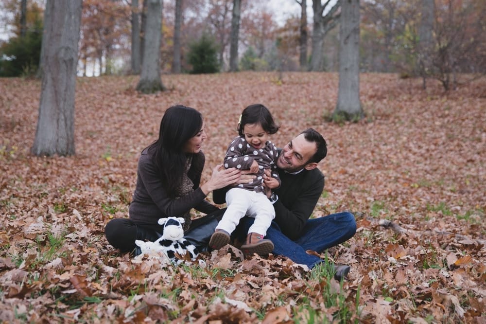 A lifestyle photograph of a family playing in the fallen leaves during their fall mini session at the Arnold Arboretum in Boston, Massachusetts