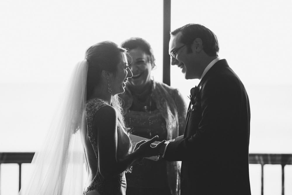 A documentary photograph of a bride and groom smiling while they exchange rings during their intimate wedding ceremony at the Oceanview of Nahant in Massachusetts