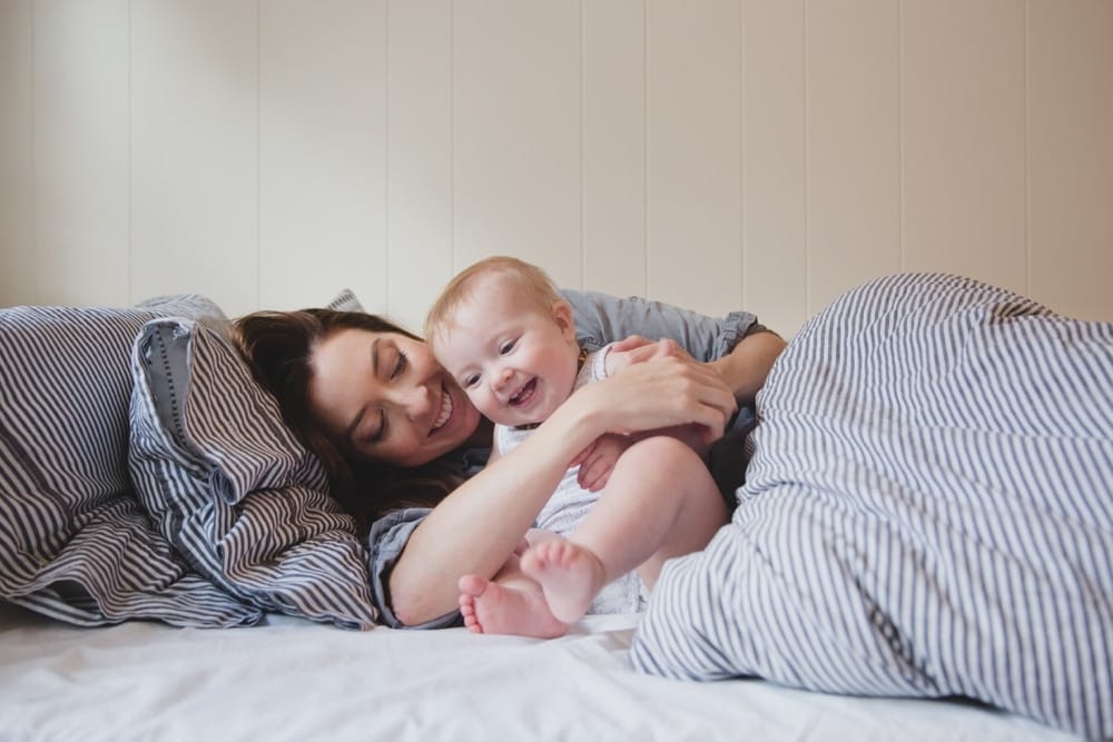 A documentary photograph of a mom and baby girl cuddling and laughing in bed during their in home lifestyle session in Boston