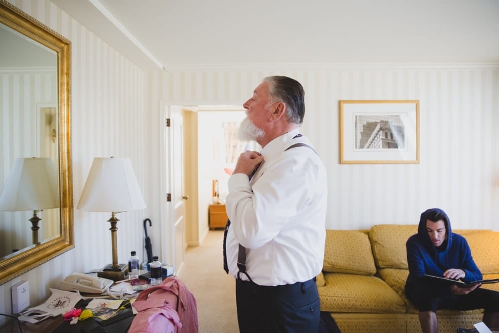 A documentary photograph of the father of the groom putting on his tie at the Langham Hotel before his son's State Room Wedding in Boston