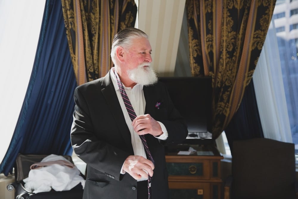 A documentary photograph of the father of the groom tying his tie at the Langham Hotel while getting ready for his son's State Room Wedding