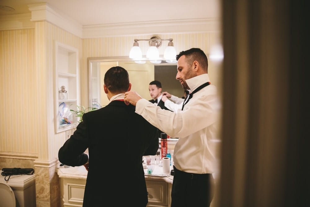 A documentary photograph of a Groomsmen straighten the Groom's bow tie in the bathroom before his Taj Boston Hotel Wedding in Masschusetts