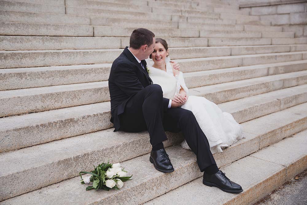 A documentary photograph of a couple laughing as they sit together on the steps of Boston Common during their wedding portrait session in Boston Public Gardens and Beacon Hill