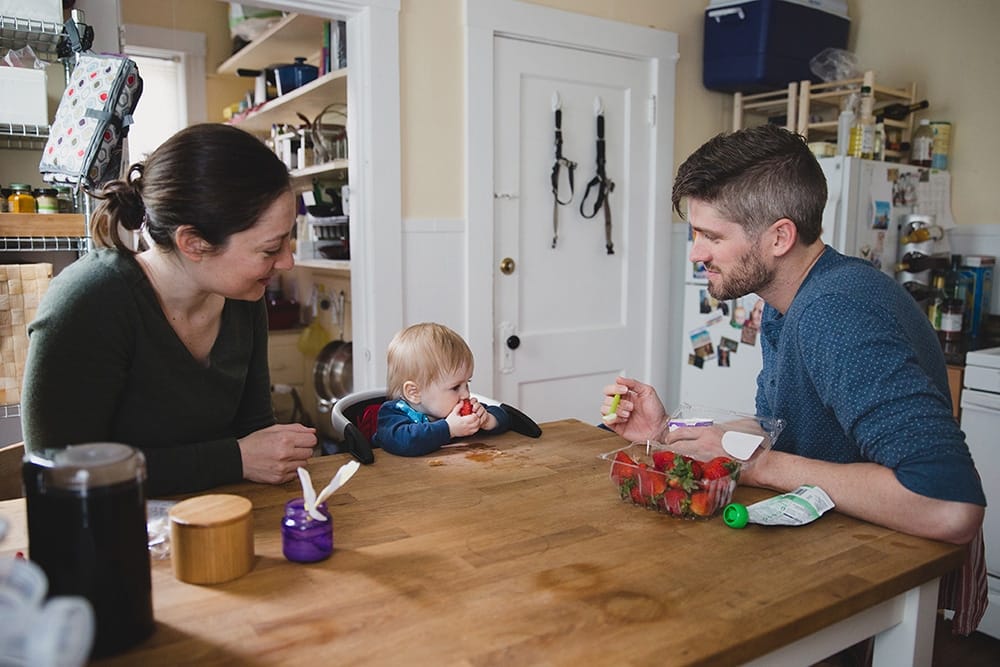 A documentary photograph of a baby boy eating a strawberry with his parents during a family session at home in Boston, Massachusetts