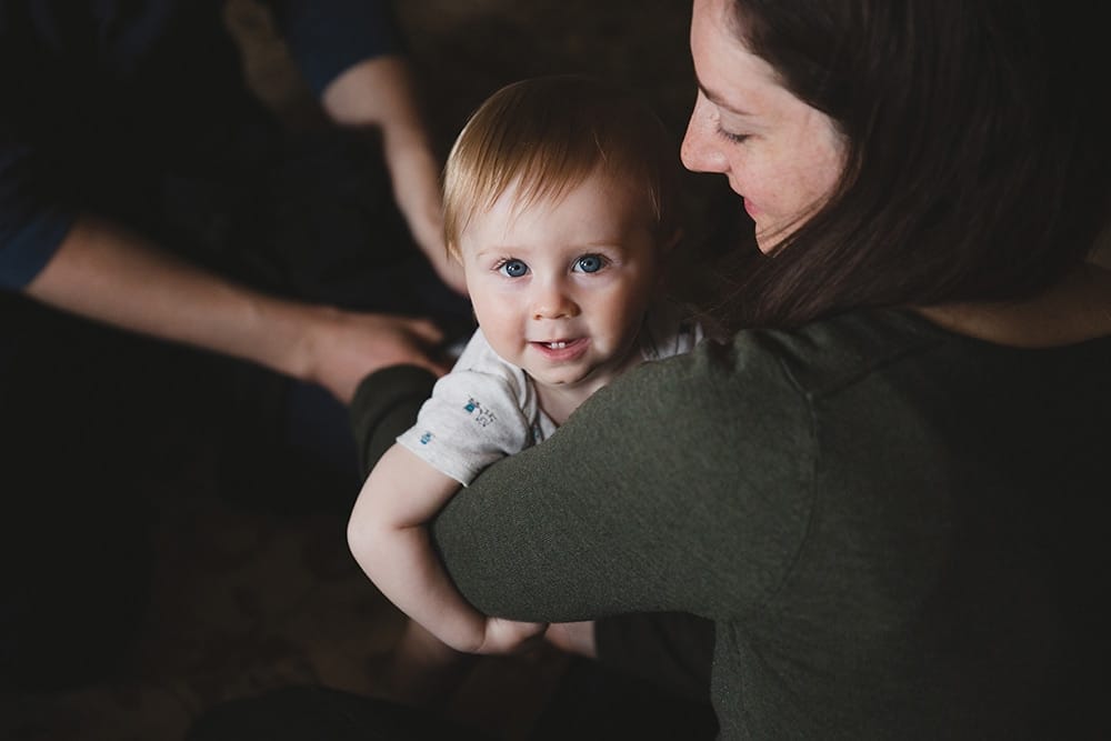A lifestyle photograph of a mother holding her son while her husbands gets him dressed during a family session at home