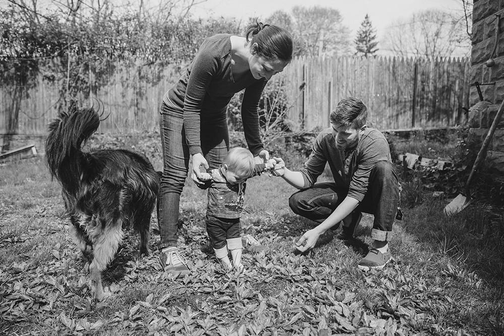 A documentary photograph of a mother and father helping their baby boy stand in the grass during a family session at home in Boston, Massachusetts