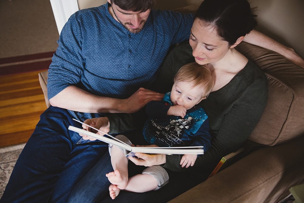 A documentary photograph of a mother and father reading a book to their baby boy during a family session at home in Boston, Massachusetts