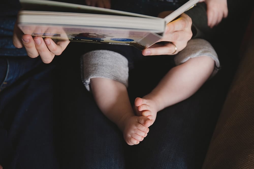 A lifestyle photograph of a mother reading a book to her baby boy during a family session at home in Boston, Massachusetts