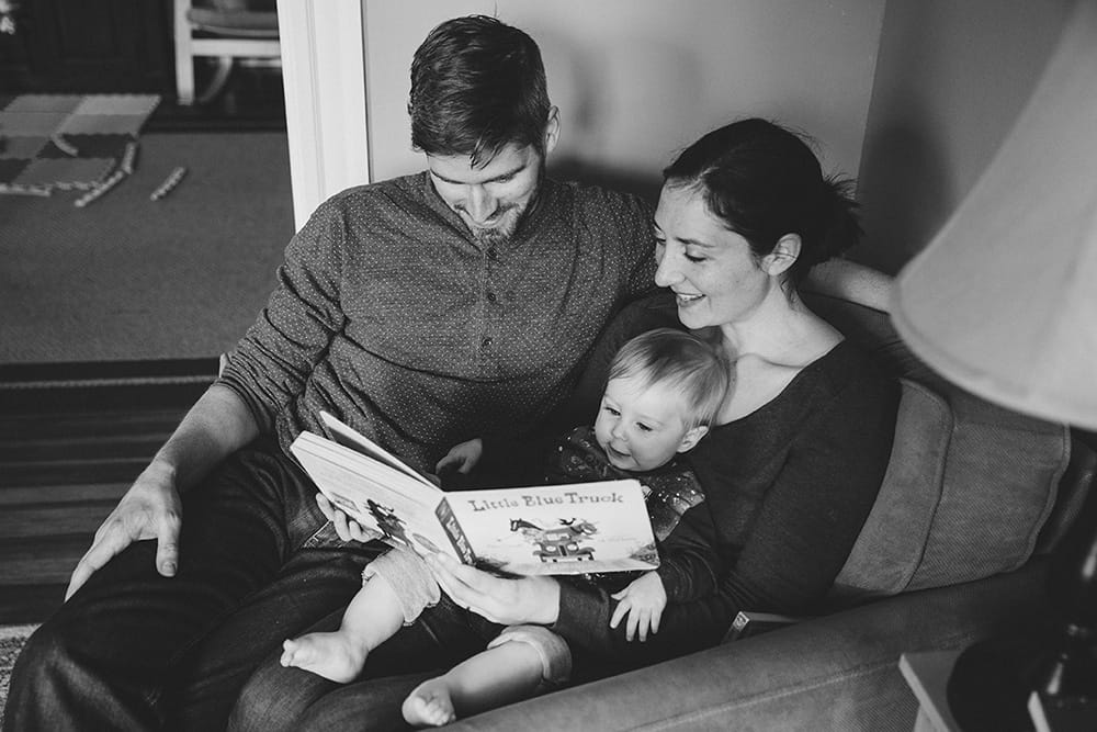 A documentary photograph of a mother and father reading the little blue truck to their baby boy during a family session at home in Boston, Massachusetts