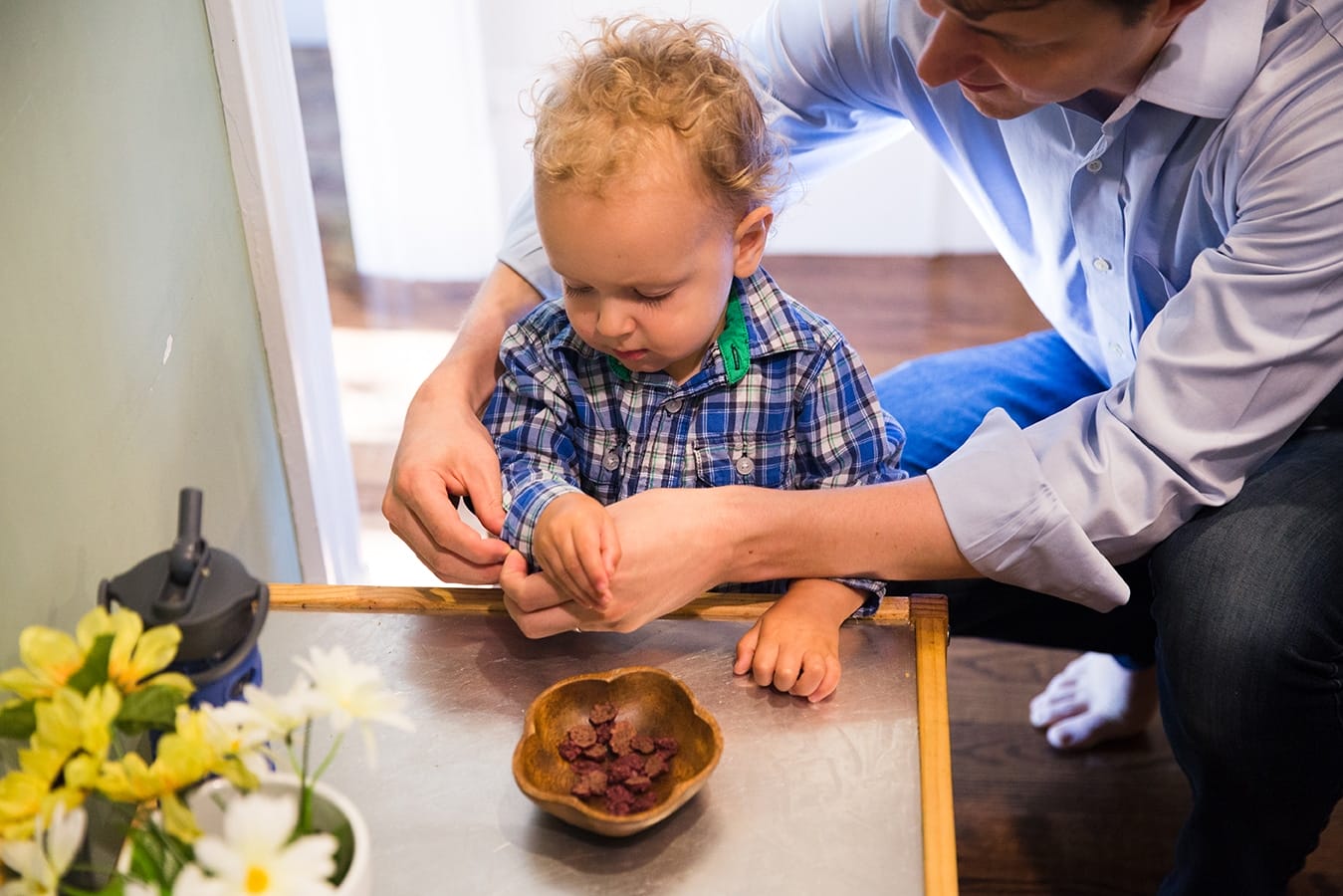 A lifestyle photograph of a Dad buttoning his toddlers sleeves as he has a snack during their in home family session in Boston