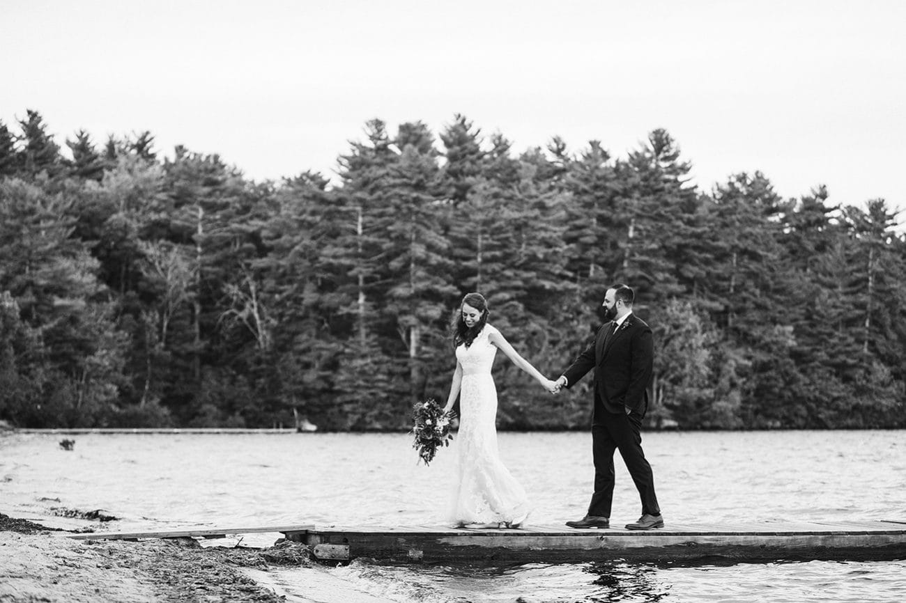 A documentary photograph of a bride and groom walking along the dock at Kingsley Pines Camp during their summer camp wedding in Maine
