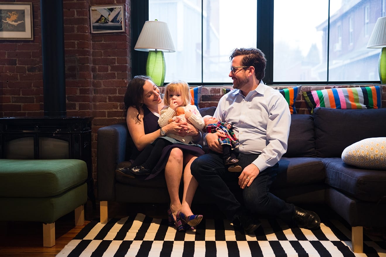 A family sit together on the couch during their in home newborn session in the Jamaica Plain neighbourhood of Boston, Massachusetts
