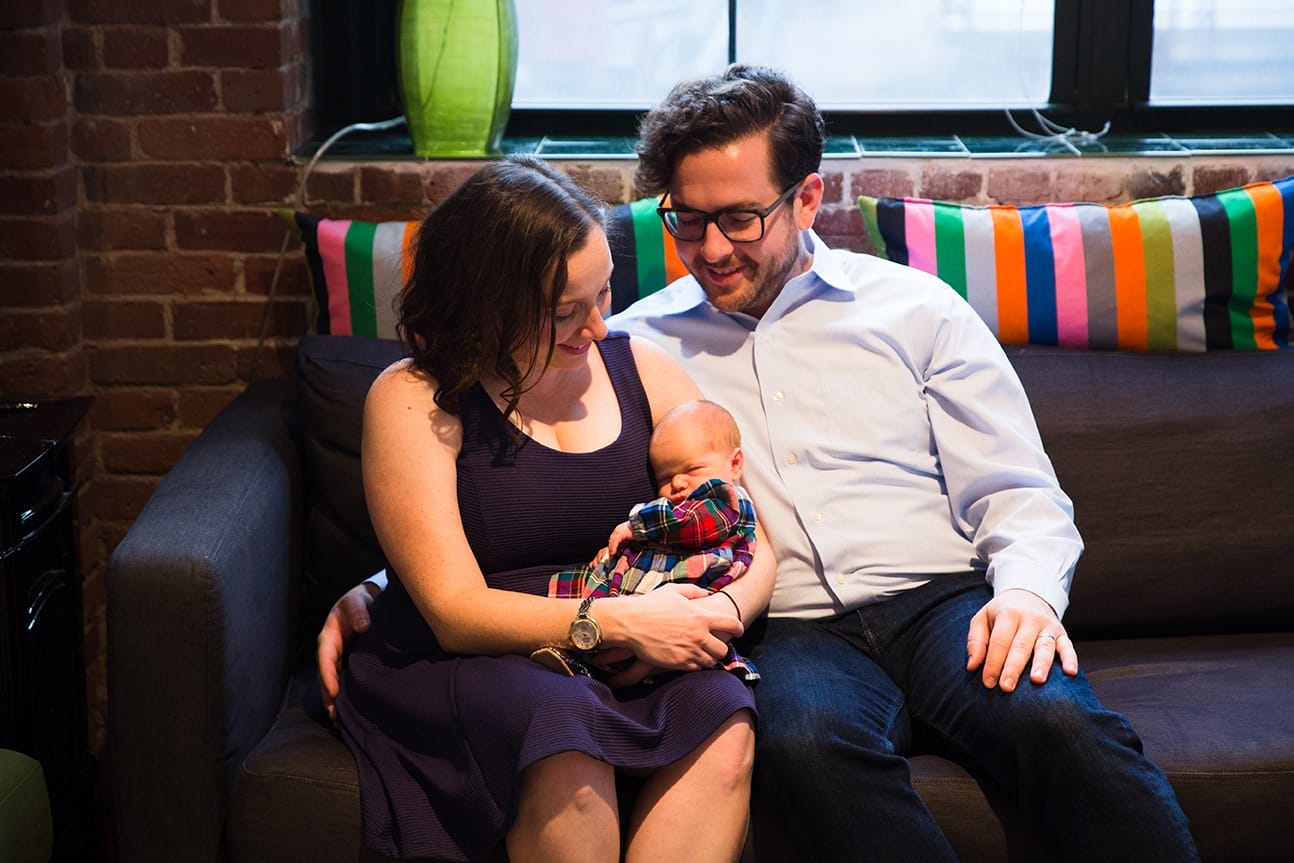 A mother and father sit on the couch with their new baby girl during an in home newborn session in the Jamaica Plain neighborhood of Boston, Massachusetts