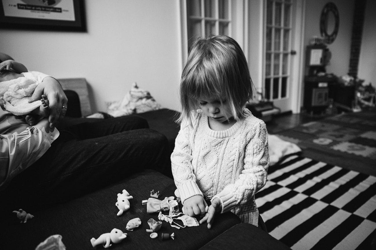 A lifestyle photograph of a toddler playing with her toys while her parents hold her new baby sister during an in home newborn session in the Jamaica Plain neighbourhood of Boston