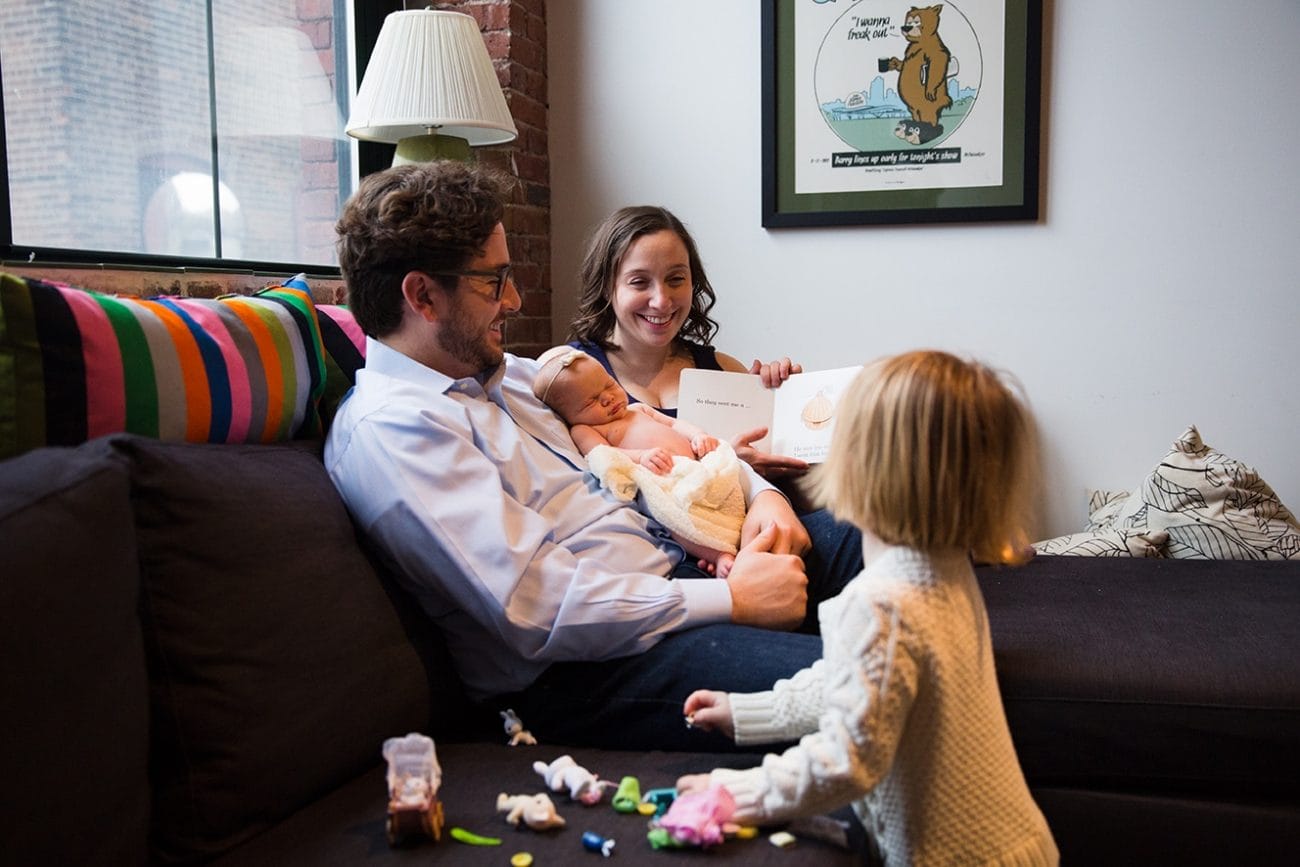 A lifestyle photograph of a mother reading her daughter a book while Dad holds their new baby girl during an in home newborn session in the Jamaica Plain neighbourhood of Boston, Massachusetts