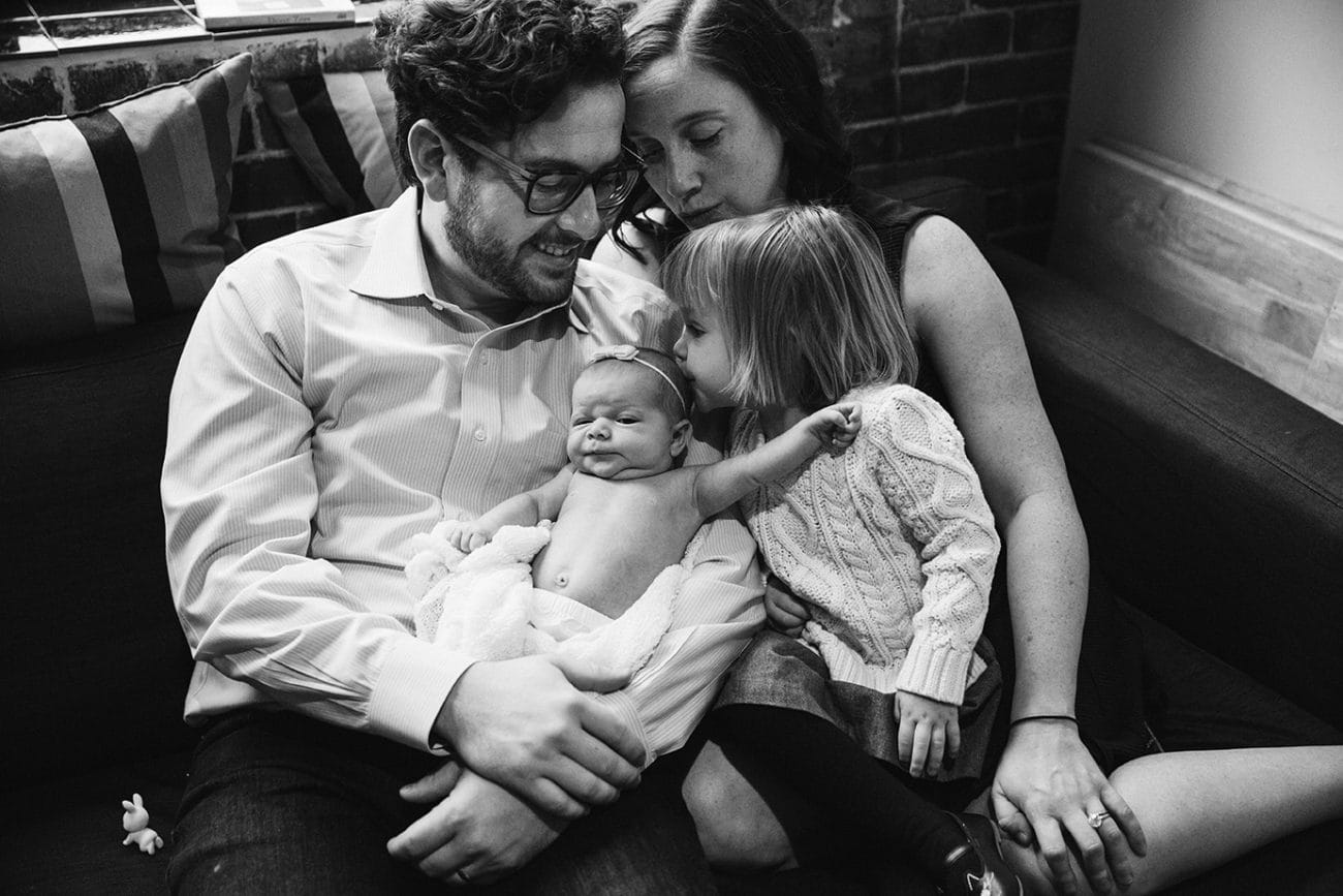 A lifestyle photograph of a sister kissing her new baby sister as they sit on the couch during their in home newborn session in the Jamaica Plain neighbourhood of Boston, Massachusetts