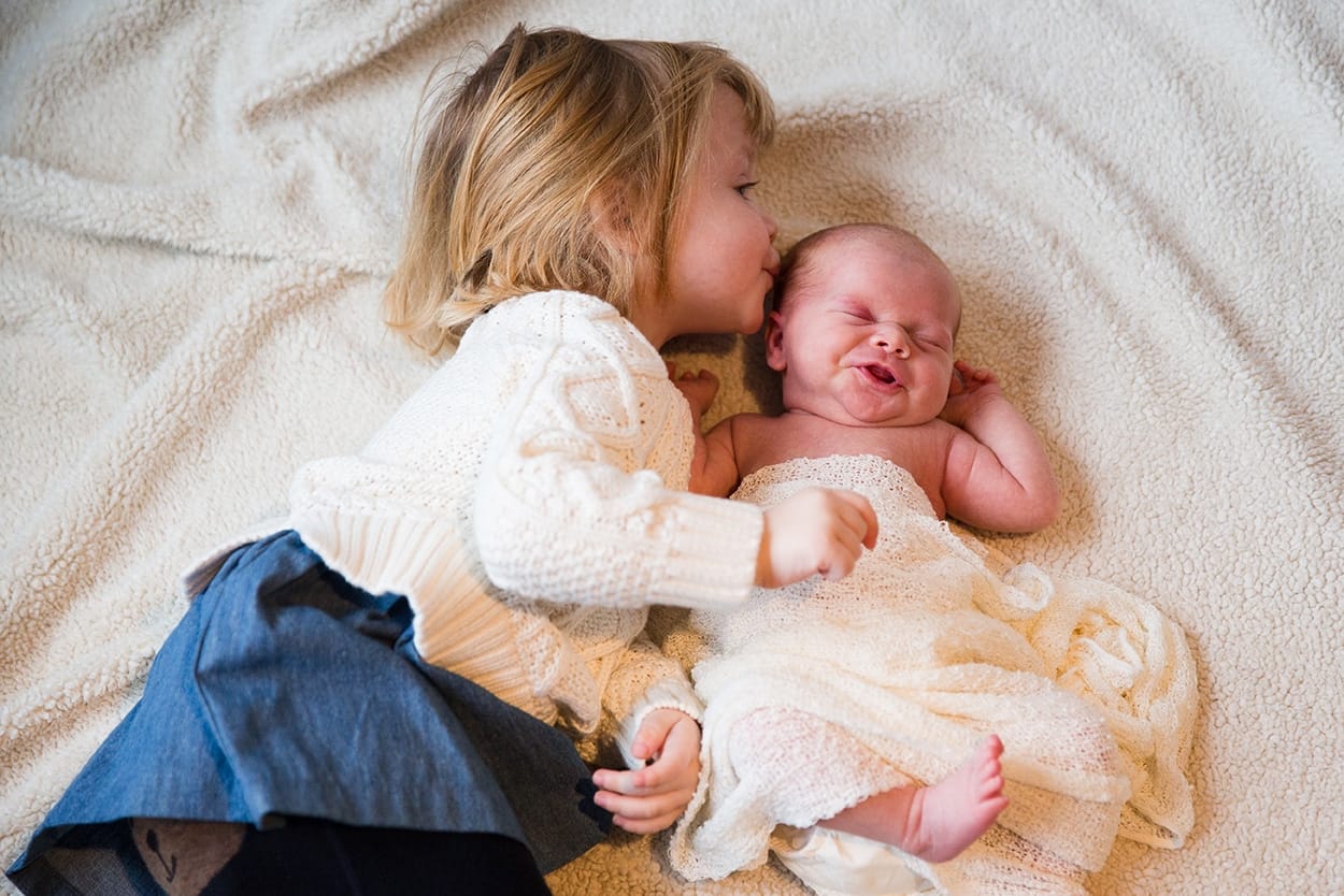 A lifestyle photograph of a toddler kissing her new baby sister during an in home newborn session in the Jamaica Plain neighbourhood of Boston, Massachusetts