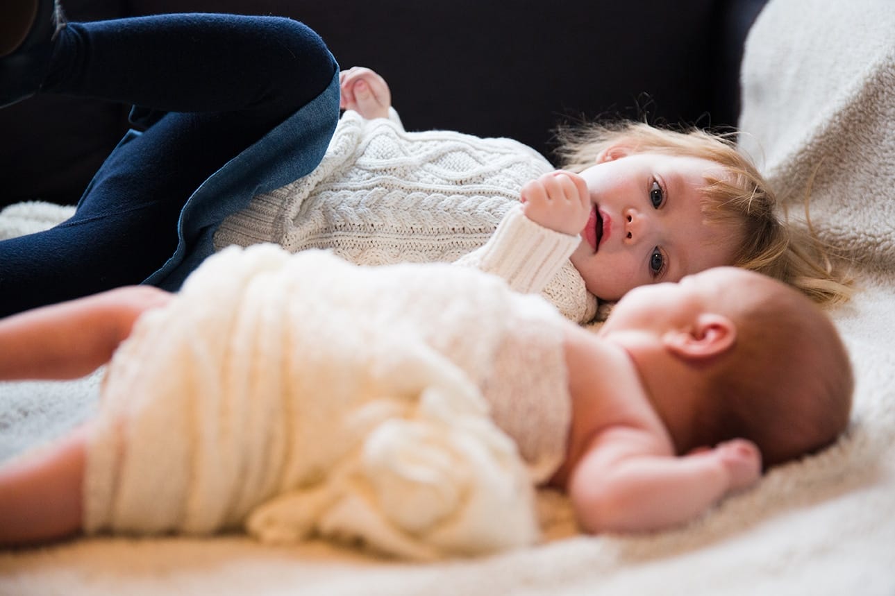 A lifestyle photograph of a toddler laying next to her new baby sister during an in home newborn session in the Jamaica Plain neighbourhood of Boston, Massachusetts
