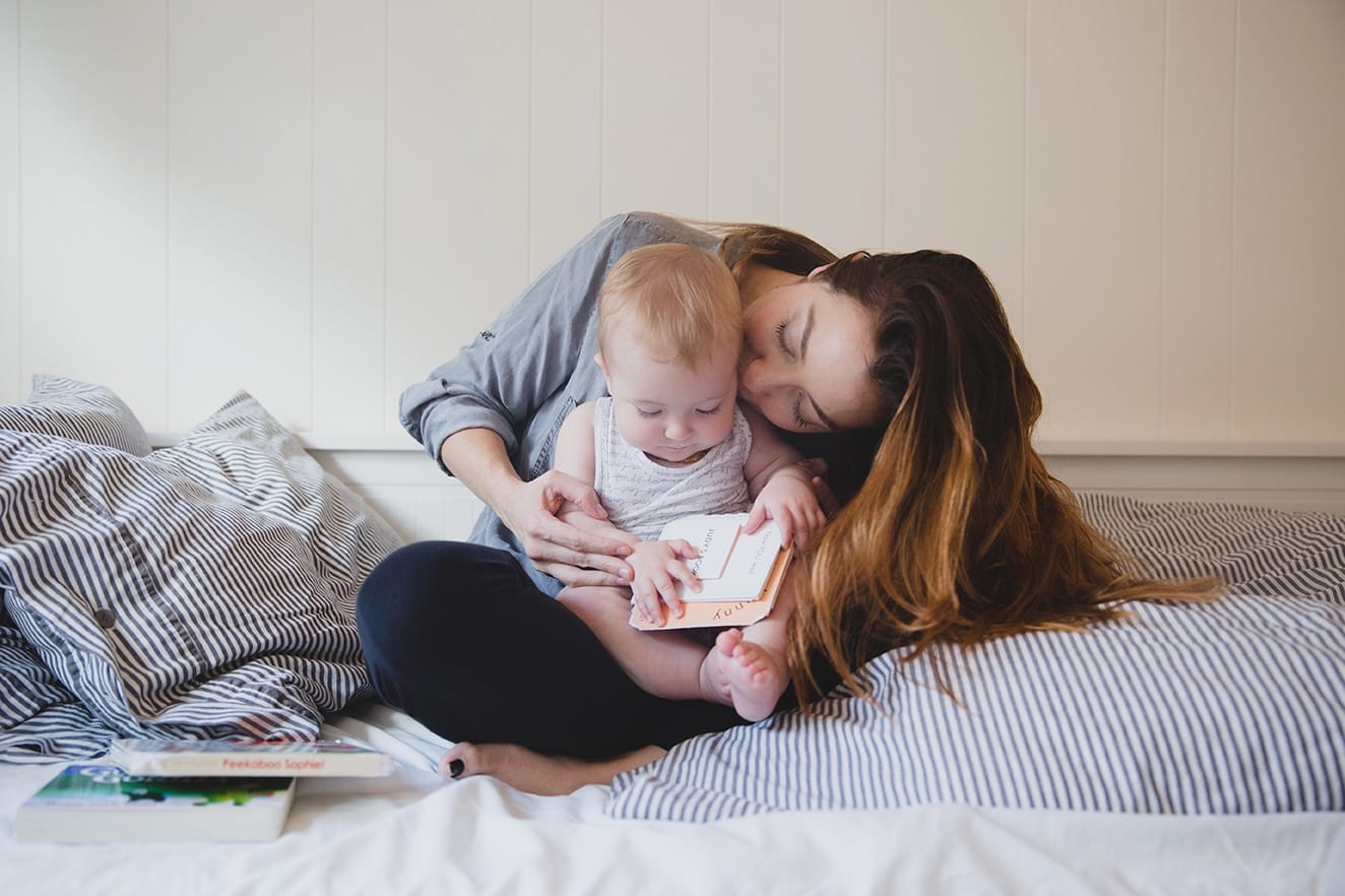 One of the best family photographs of 2016 where a mom kisses her daughter while they read books together on the bed during an in home family session