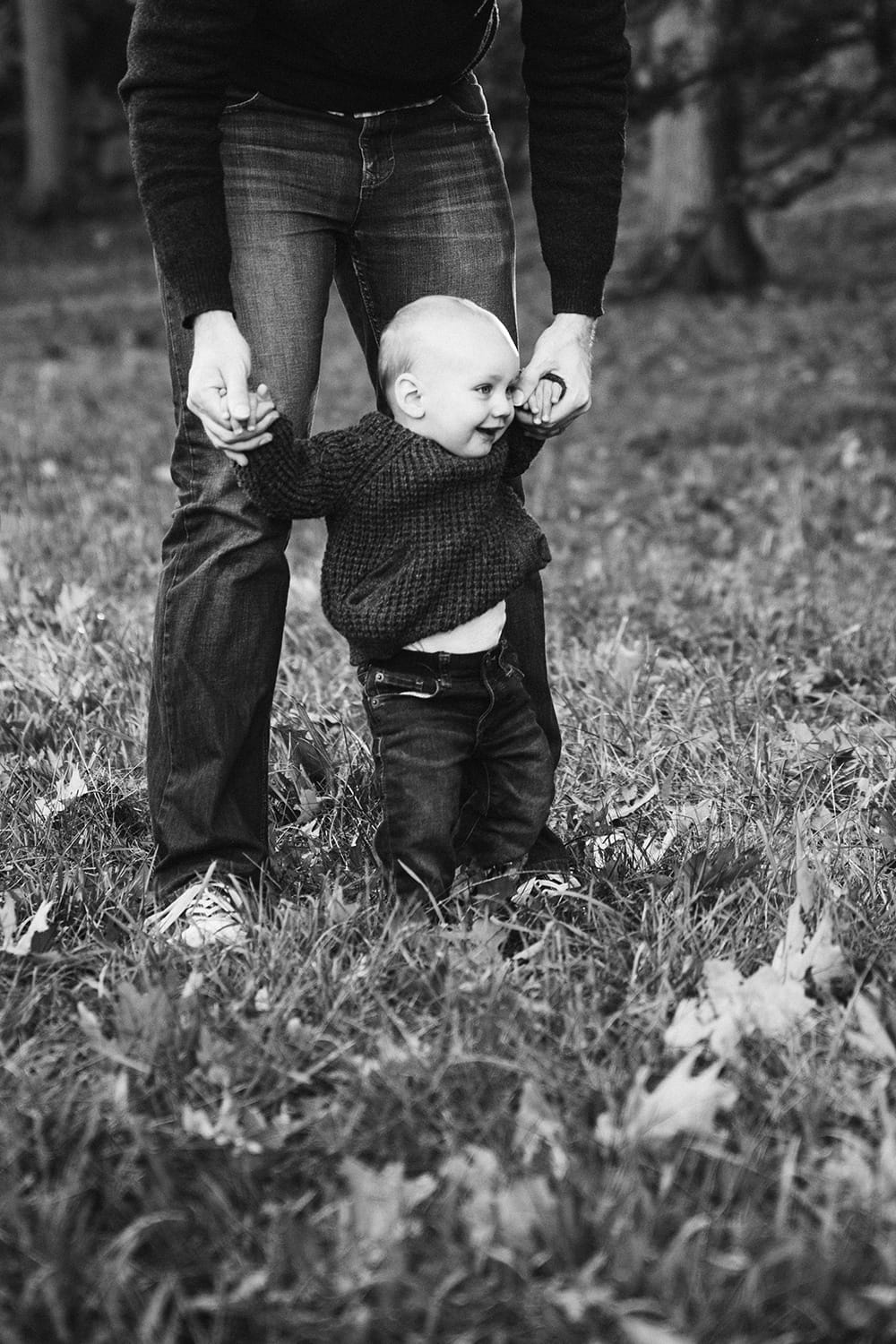 A documentary photograph of a father helping his baby boy walk during their lifestyle family photo session in the Arnold Arboretum