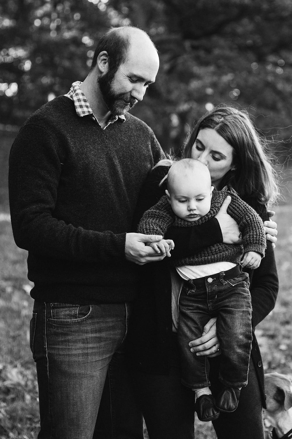 A lifestyle portrait of a mother and father kissing and holding their baby boy during their Arboretum Family Session in Boston, Massachusetts