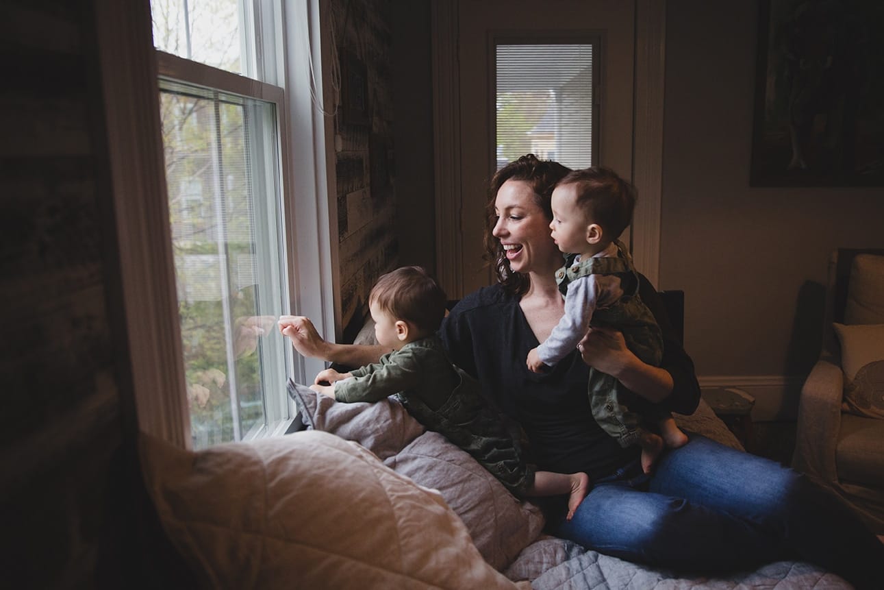 A documentary photograph of a mom looking out the window with her twin boys during an in home family session in Boston, Massachusetts