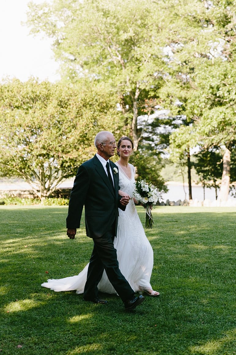 A documentary photograph of a bride walking up the aisle with her father during a Moraine Farm wedding in Beverly, Massachusetts